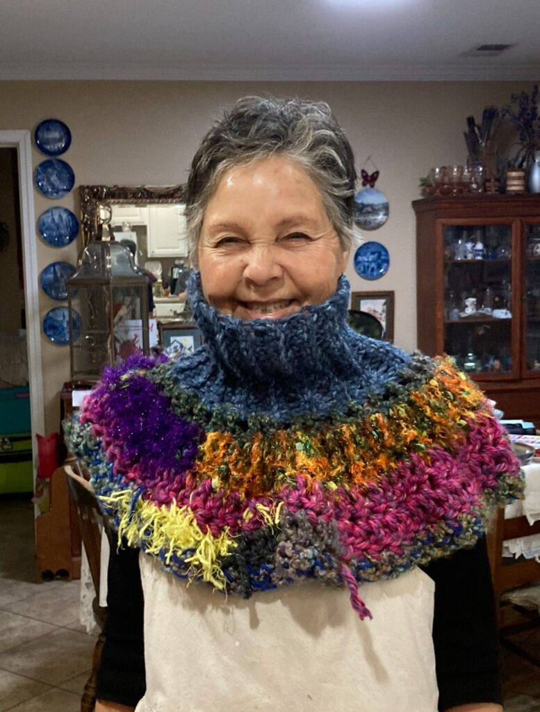A mature women stands in a vintage style kitchen with a large smile and greying hair wearing a colorful patchwork of crochet yarn.